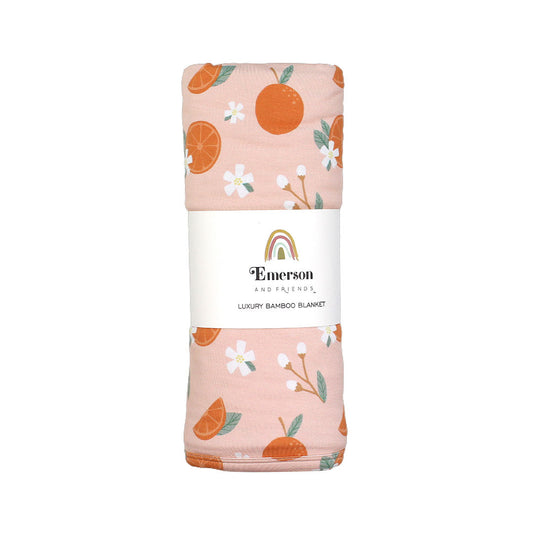 the "freshly squeezed" bamboo blanket. the "freshly squeezed" print has an assortment of full and half cut oranges scattered around. there is also flower heads and flower stems that intermingle within the print. this is all space out around a pink background space. 