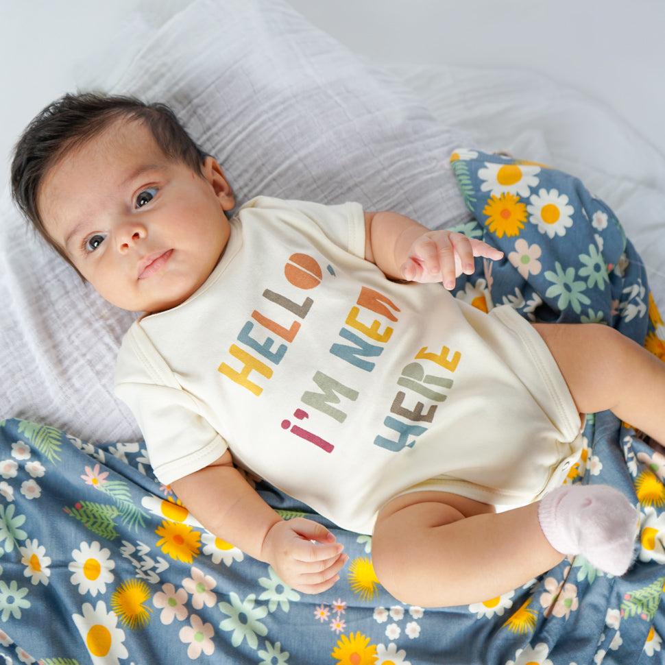 small baby lays on their back. the babu is laying on the "blue daisy" bamboo blanket. the baby wears the "hello, i'm new here" bamboo onesie. the "blue daisy" print is an arrangement of tons of different, colorful flowers scattered around a dark blue print. 