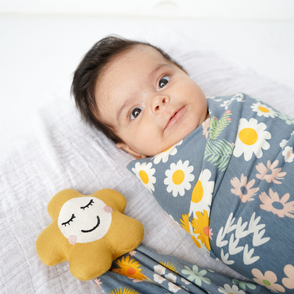 little baby stares wide eye into the camera. the baby is wrapped in the "blue daisy" blanket. the lovey is placed to the baby. the "blue daisy" print is an arrangement of tons of different, colorful flowers scattered around a dark blue print. 