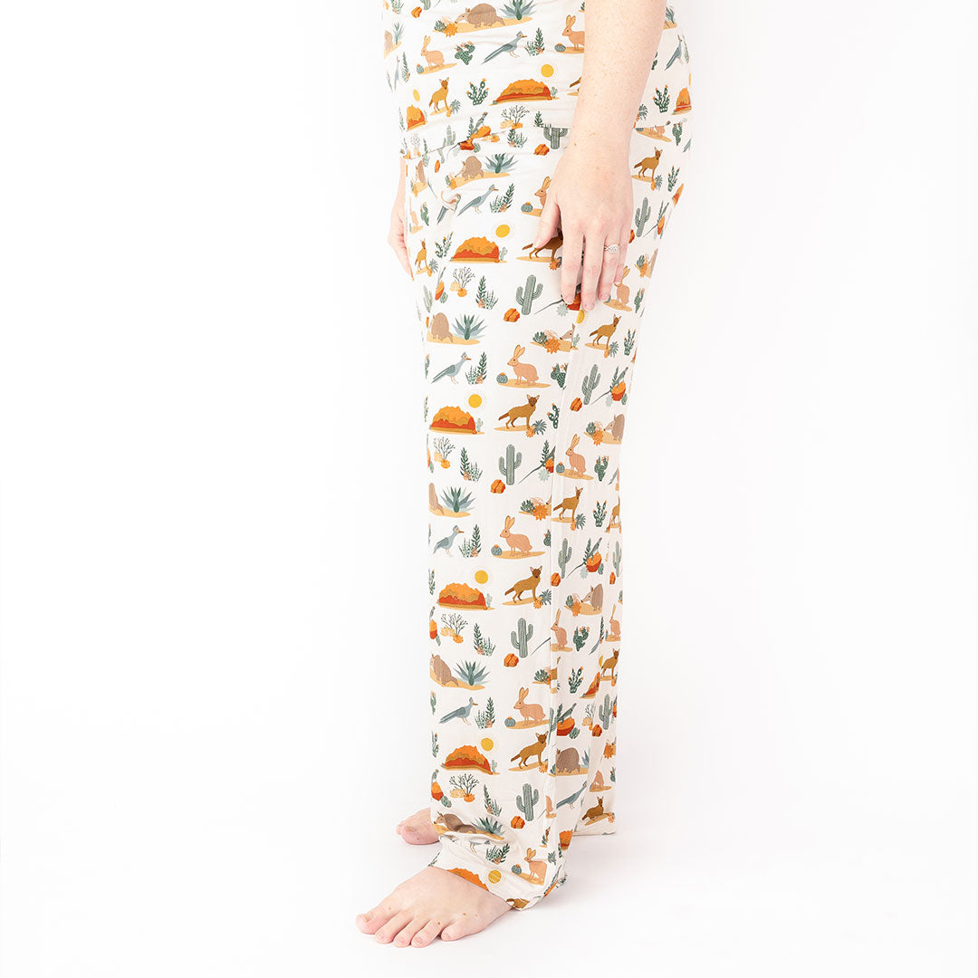 person stands in their "desert friends" relaxed pants. the "desert friends" print is a scattered pattern of armadillo, cacti, other desert plants, rocks, desert foxes, lizards, and desert birds. this is all on a beige background. 