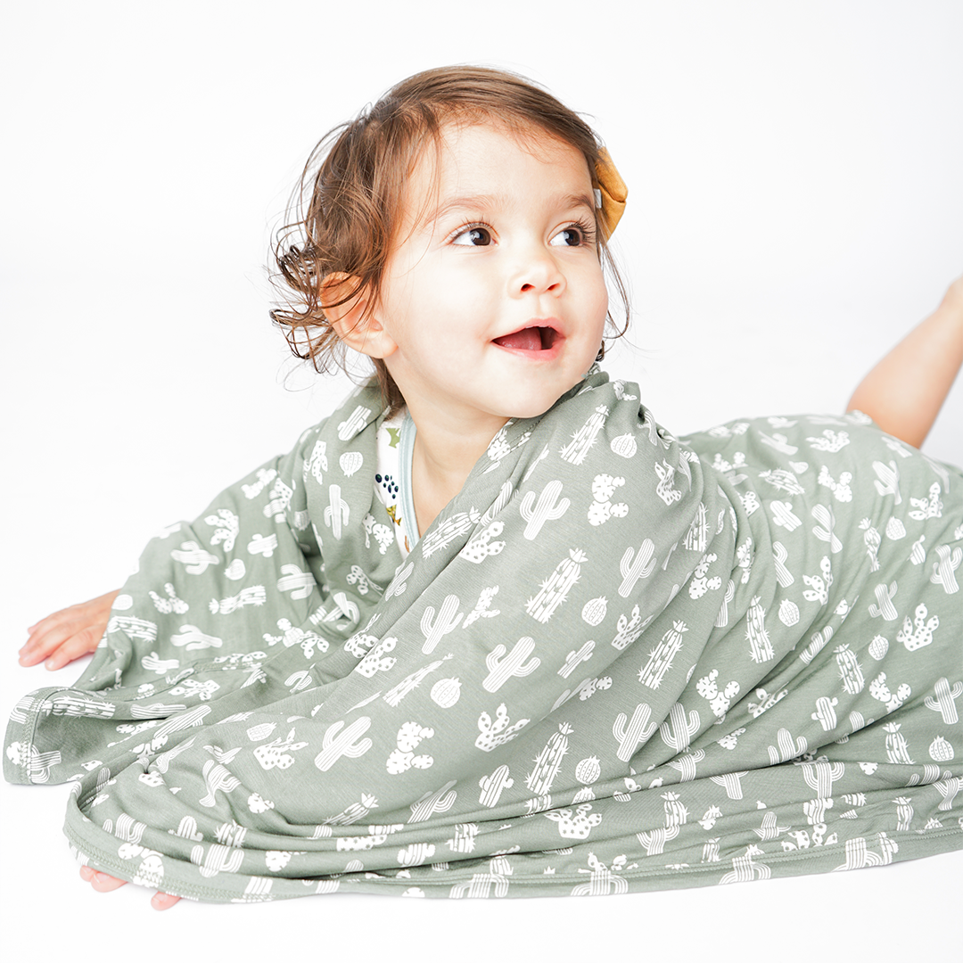 little baby lays on their stomach and wraps themself in the "stay sharp" bamboo blanket. the "stay sharp" print is a variety of different white cacti on a greyish/green background. 