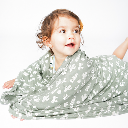 little baby lays on their stomach and wraps themself in the "stay sharp" bamboo blanket. the "stay sharp" print is a variety of different white cacti on a greyish/green background. 