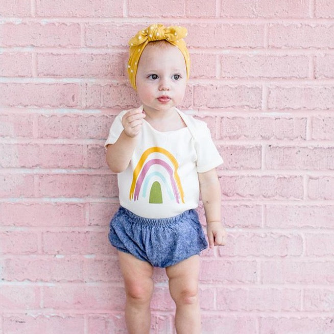 a small girl up against a pink wall. she is wearing the "rainbow" baby cotton onesie. a white onesie with a boho yellow, pink, teal, olive green rainbow. 