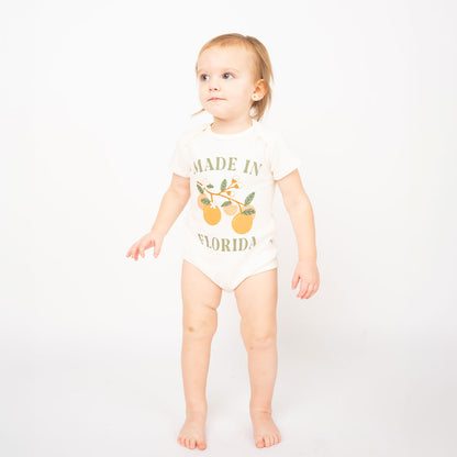 the baby is standing up and looking away from the camera. they are wearing the "made in Florida" onesie. this cotton onesie has the phrase "made in Florida" in green print on the onesie. there is also a branch of oranges on it too. 