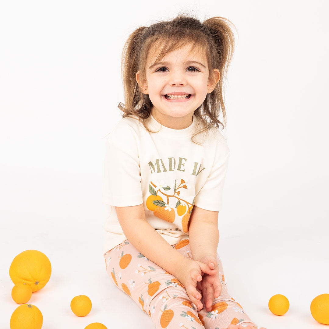 cute toddler girl sits with her legs long and her hands in her lap. she has oranges scattered around her. she is wearing the cotton "made in florida" t-shirt and the "freshly squeezed" pajama bottoms.