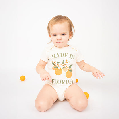 a baby sitting on their knees with oranges scattered around them. they are wearing the "made in Florida" cotton onesie. this cotton onesie has the phrase "made in Florida" in green print on the onesie. there is also a branch of oranges on it too. 