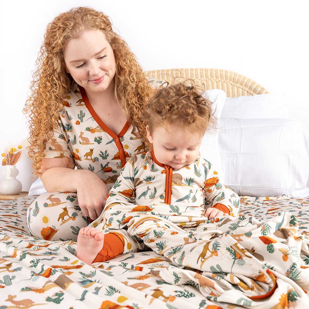 a mom and baby sit on a bed. the bed is covered with the "desert friends" bamboo blanket. the mom and baby are matching in their "desert friends" attire. the mom in the women's shirt and the joggers, the baby in the convertible.the "desert friends" print is a scattered pattern of armadillo, cacti, other desert plants, rocks, desert foxes, lizards, and desert birds. this is all on a beige background. 