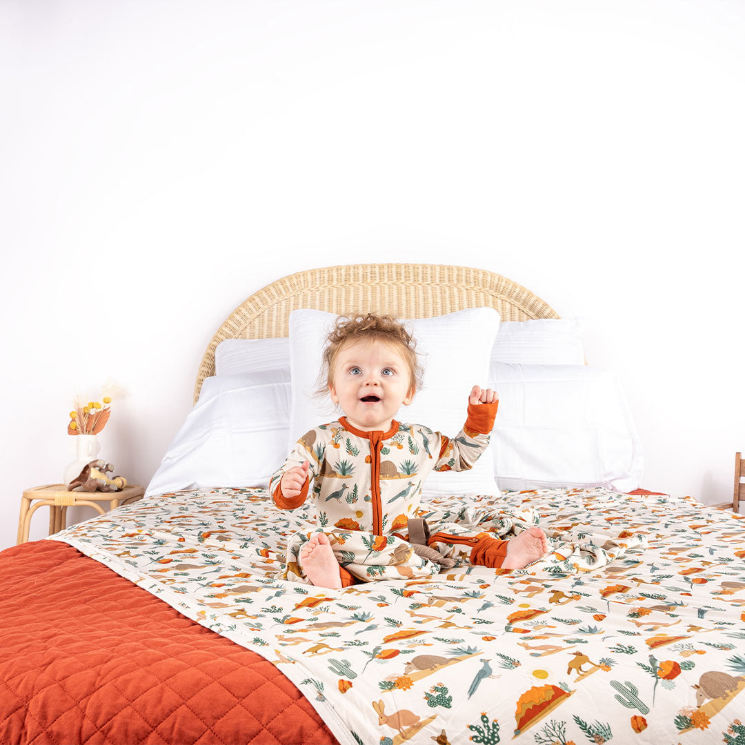 a baby sit on a bed with their hands in the air. the baby is wearing the desert friends convertible. the bed has the twin sized blanket draped over it. the "desert friends" print is a scattered pattern of armadillo, cacti, other desert plants, rocks, desert foxes, lizards, and desert birds. this is all on a beige background. 