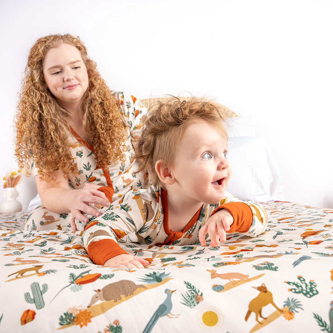 mom and baby play on a bed. the bed has the "desert friends" twin blanket drapped over it. the baby is laying on it's tummy, laughing, wearing the "desert friends" convertible. the "desert friends" print is a scattered pattern of armadillo, cacti, other desert plants, rocks, desert foxes, lizards, and desert birds. this is all on a beige background. 