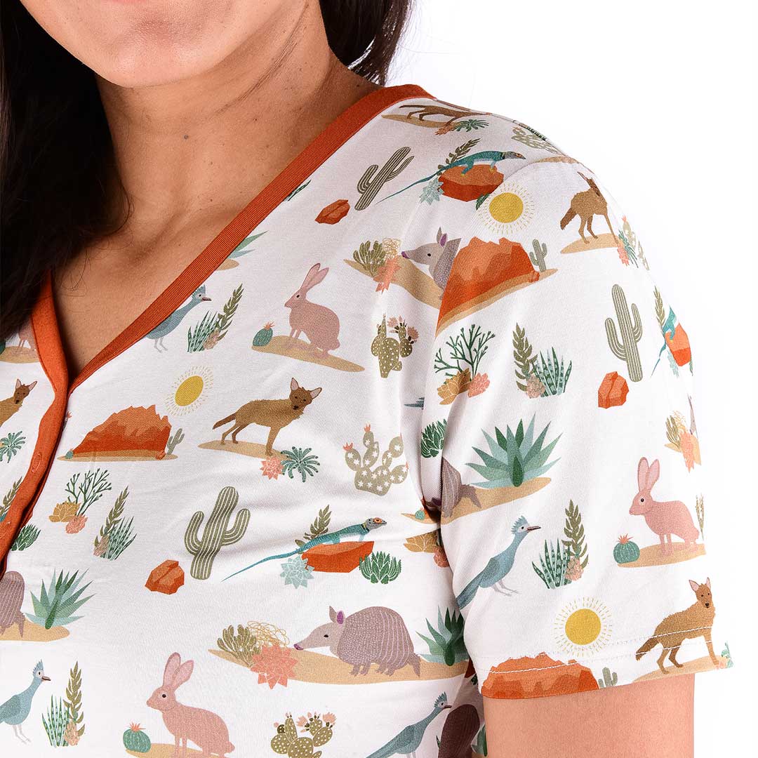 an up close version of the "desert friends" women's top. it shows the shoulder of a women. the "desert friends" print is a scattered pattern of armadillo, cacti, other desert plants, rocks, desert foxes, lizards, and desert birds. this is all on a beige background. 