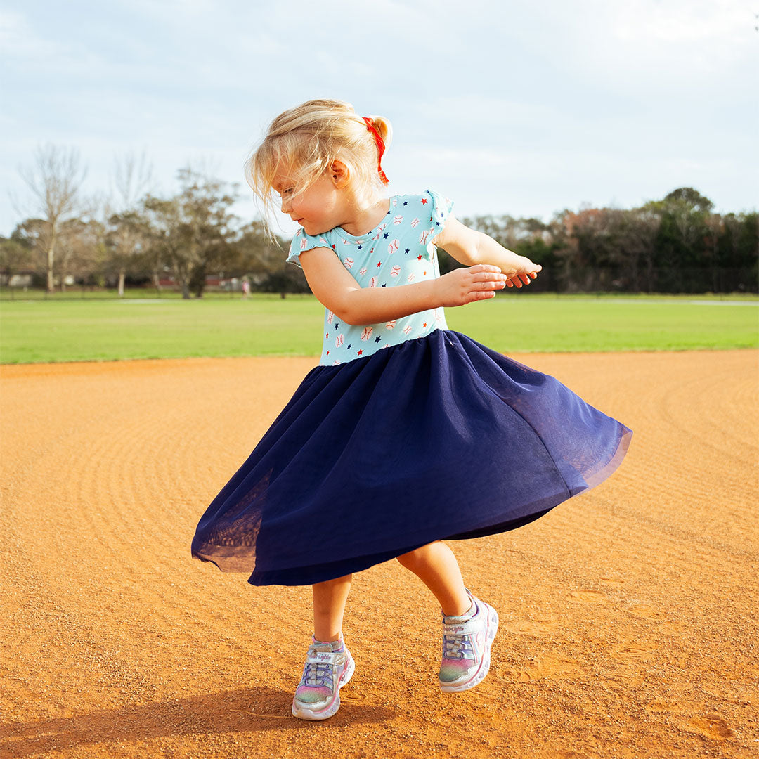 cute toddler girl is spinning around on the baseball field. she is wearing the "baseball buddies" twirl dress. the "baseball buddies" print is a combination of baseballs, red stars, and blue stars scattered across a teal background. 