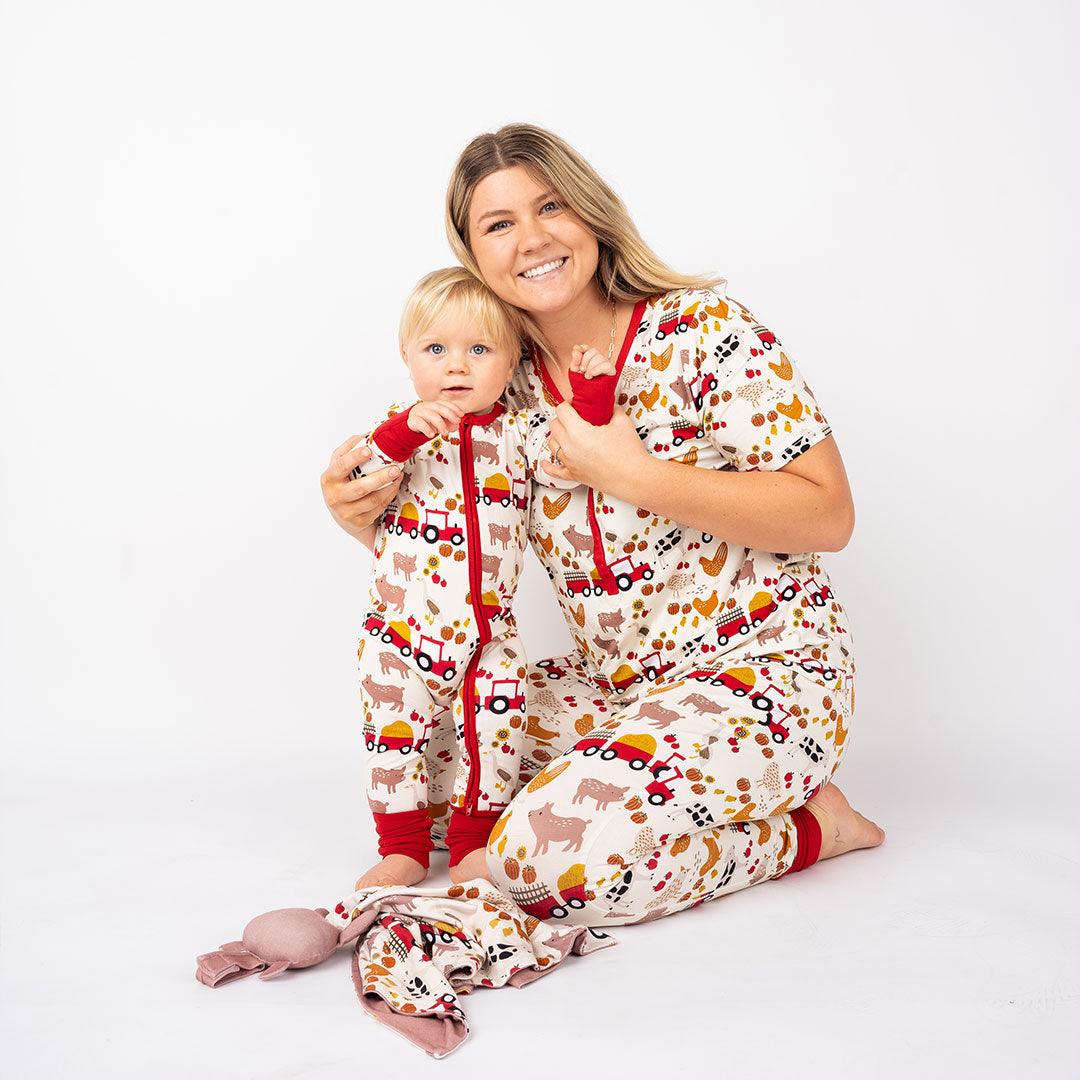 a mom sits on the floor with her child. they both smile for the camera, wearing the "farm friends" prints in the women's joggers, top, and the convertible. the "farm friends" print is a collection of chickens, roosters, pigs, bails of hay, tractors, chicks, apples, and pumpkins. it's a white background and flashed of reds, yellows, and oranges, to bring out the farm aesthetic.