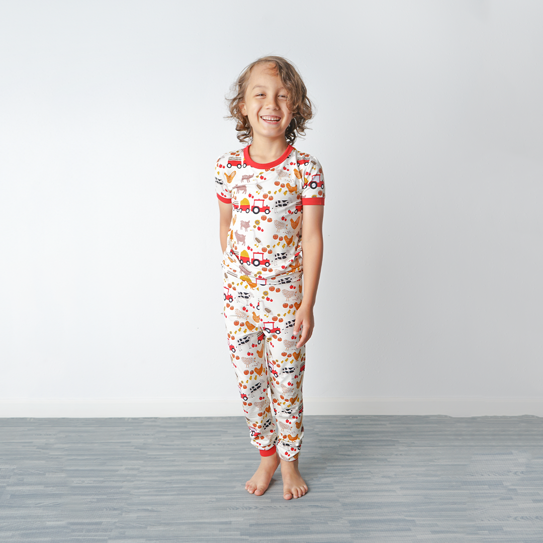 a girl stands and laughs at the camera. she is wearing the "farm friends" matching pajama set. the "farm friends" print is a collection of chickens, roosters, pigs, bails of hay, tractors, chicks, apples, and pumpkins. it's a white background and flashed of reds, yellows, and oranges, to bring out the farm aesthetic.