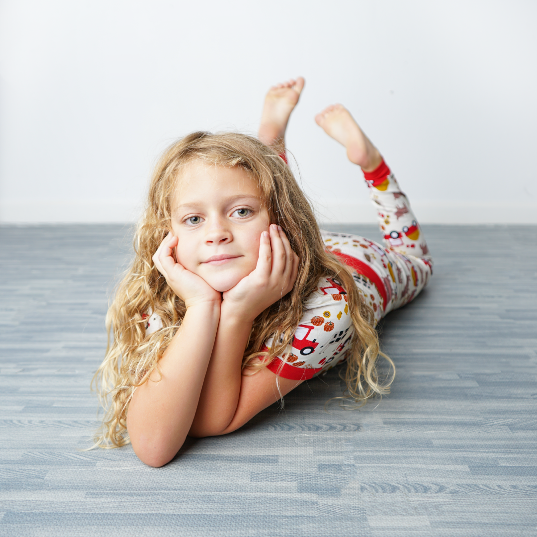 a young girl lays on her stomach with her hands cupping her face. her feet are in the air. she wears the "farm friends" matching pajama set. the "farm friends" print is a collection of chickens, roosters, pigs, bails of hay, tractors, chicks, apples, and pumpkins. it's a white background and flashed of reds, yellows, and oranges, to bring out the farm aesthetic.