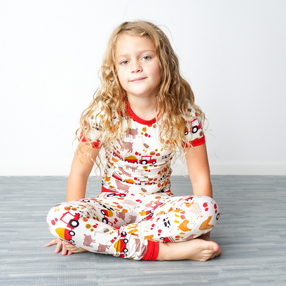 a young girl sits criss-cross on the floor. she is wearing the "farm friends" matching pajama set. the "farm friends" print is a collection of chickens, roosters, pigs, bails of hay, tractors, chicks, apples, and pumpkins. it's a white background and flashed of reds, yellows, and oranges, to bring out the farm aesthetic.