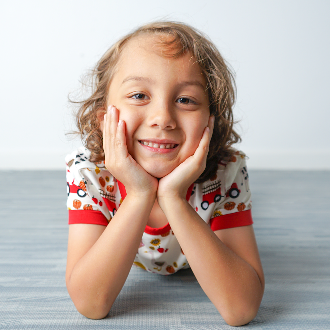 young girl lays on her stomach and smiles with her hands cupping her face. she wears the "farm friends" matching pajama set. the "farm friends" print is a collection of chickens, roosters, pigs, bails of hay, tractors, chicks, apples, and pumpkins. it's a white background and flashed of reds, yellows, and oranges, to bring out the farm aesthetic.