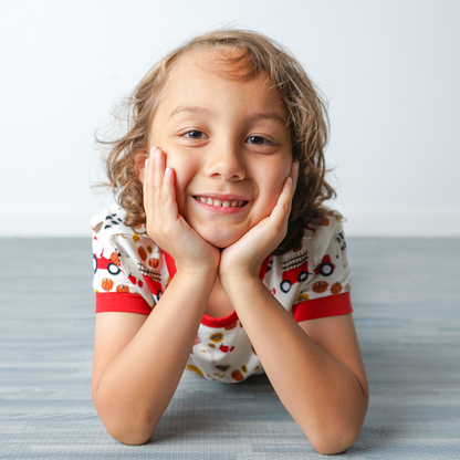 young girl lays on her stomach and smiles with her hands cupping her face. she wears the "farm friends" matching pajama set. the "farm friends" print is a collection of chickens, roosters, pigs, bails of hay, tractors, chicks, apples, and pumpkins. it's a white background and flashed of reds, yellows, and oranges, to bring out the farm aesthetic.