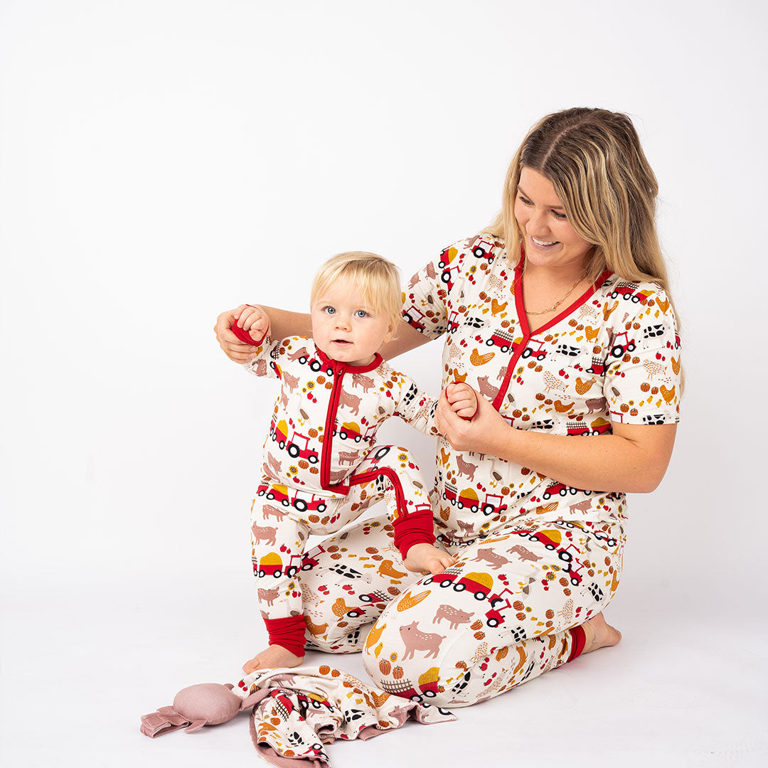 a mom sits on the floor on her knees, she wears the "farm friends" women's top. she holds her baby up while he tries to stand on her knees. the baby wears the "farm friends" convertible. the "farm friends" print is a collection of chickens, roosters, pigs, bails of hay, tractors, chicks, apples, and pumpkins. it's a white background and flashed of reds, yellows, and oranges, to bring out the farm aesthetic.