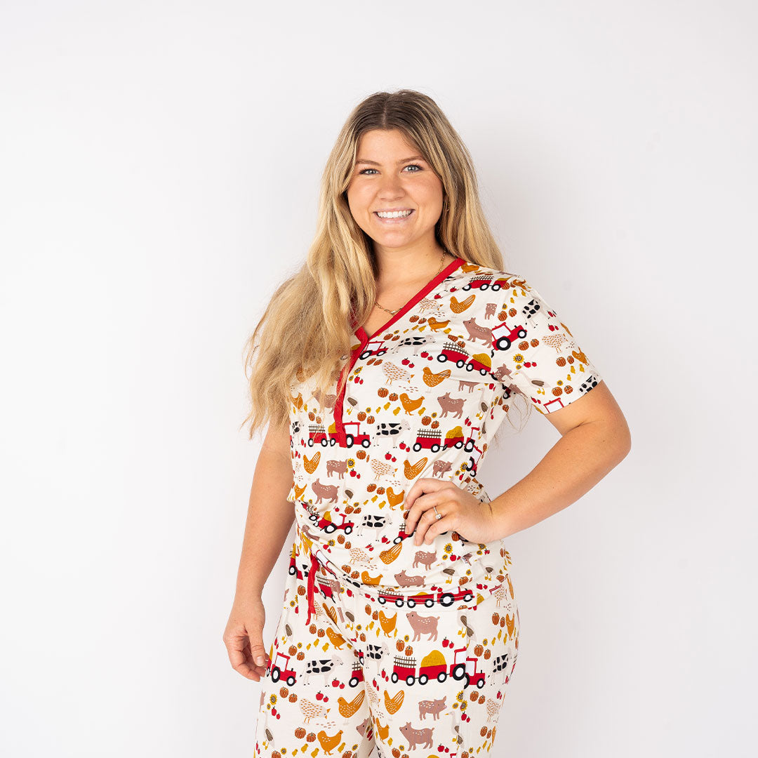 a women poses with her hand on her hip. you can see her wearing the "farm friends" women's top. the "farm friends" print is a collection of chickens, roosters, pigs, bails of hay, tractors, chicks, apples, and pumpkins. it's a white background and flashed of reds, yellows, and oranges, to bring out the farm aesthetic.