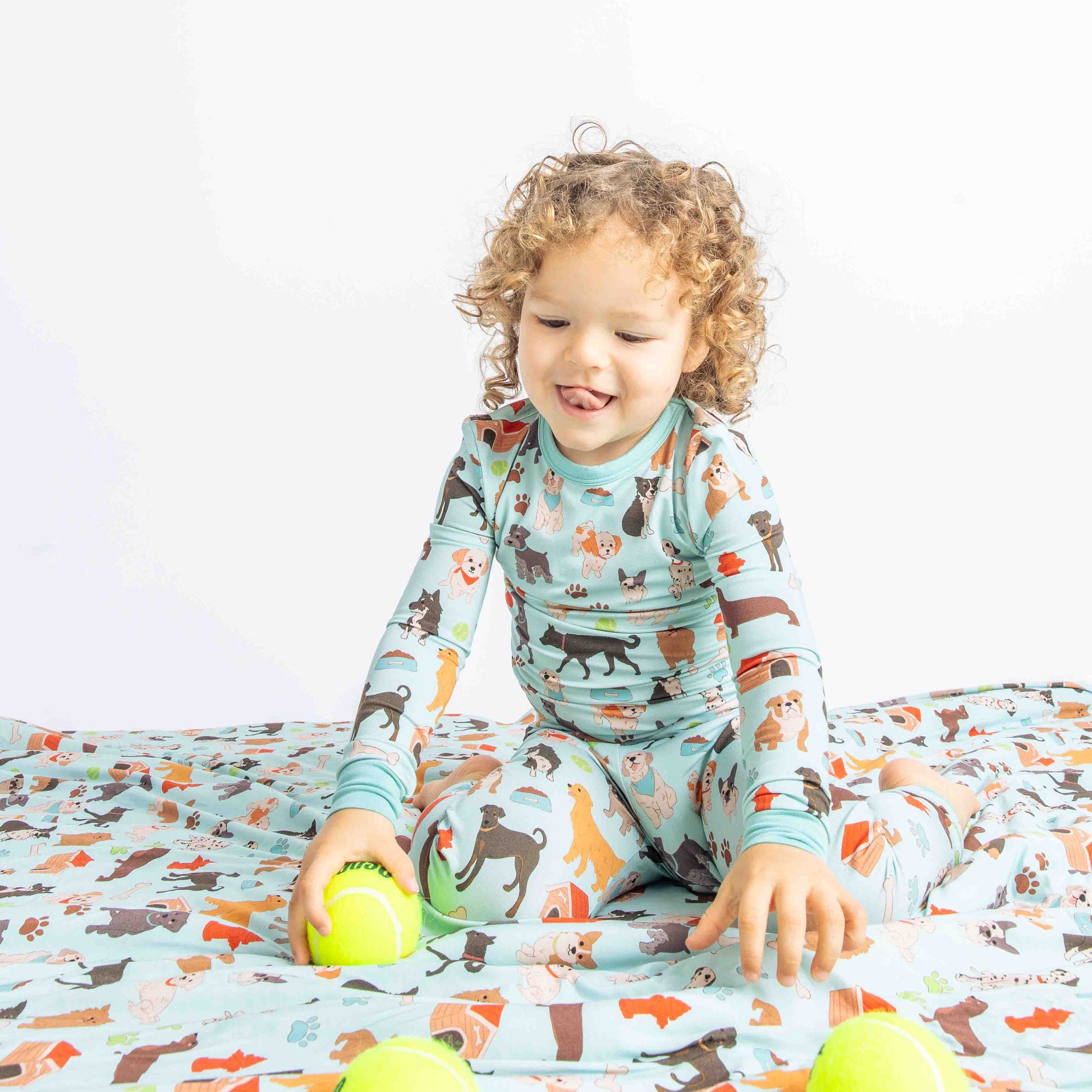 child in the "dogs bark" matching 2-piece pajama set sits on the "dogs bark" fleece blanket. he plays with tennis balls. the "dogs bark" print is a mix of various types of dogs, dog houses, dog bones, paw prints, dog food, and tennis balls scattered around a light blue background.