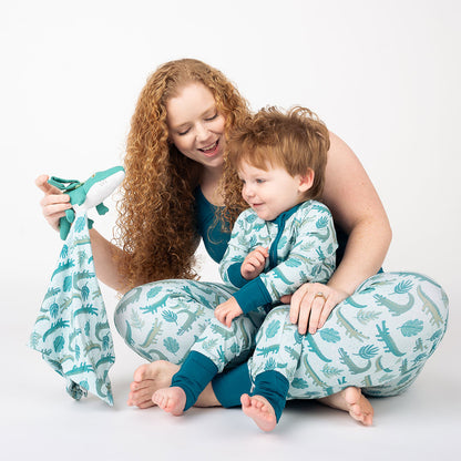 A mother with her son in her lap. they are both sporting different attire in the "later gator" prints. they are playing with the "later gator" lovey. the "later gator" print has a mix of light and dark green alligators, leaves, and white dots scattered on a teal colored background. 