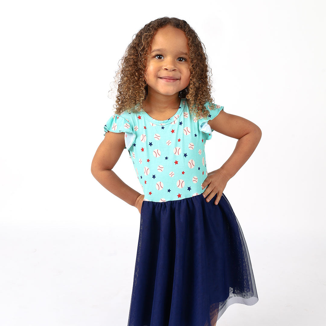 cute toddler girl poses in her "baseball buddies" twirl dress. the "baseball buddies" print is a combination of baseballs, red stars, and blue stars scattered across a teal background. 