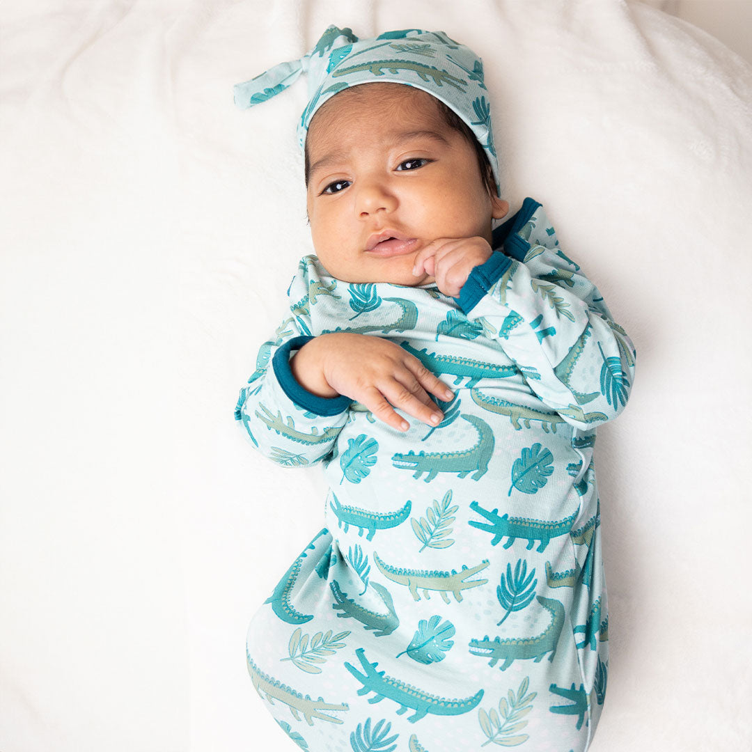 little baby in his matching gown and hat. the "later gator" print has a mix of light and dark green alligators, leaves, and white dots scattered on a teal colored background. 