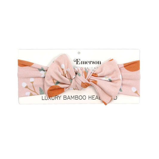 the "freshly squeezed" baby bamboo headband. the "freshly squeezed" print has an assortment of full and half cut oranges scattered around. there is also flower heads and flower stems that intermingle within the print. this is all space out around a pink background space. 