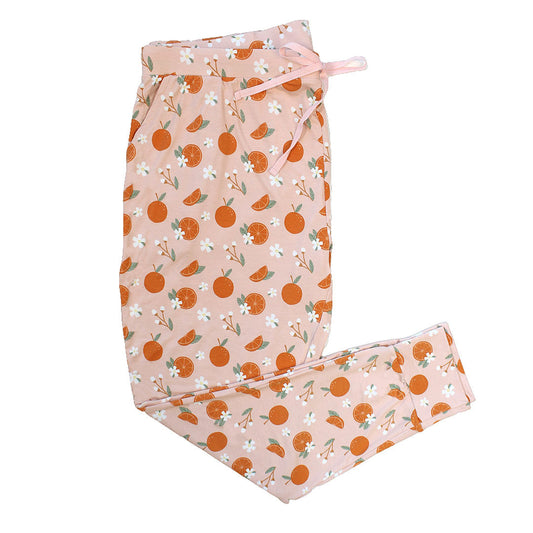 the "freshly squeezed" women's jogger pants. the "freshly squeezed" print has an assortment of full and half cut oranges scattered around. there is also flower heads and flower stems that intermingle within the print. this is all space out around a pink background space. 