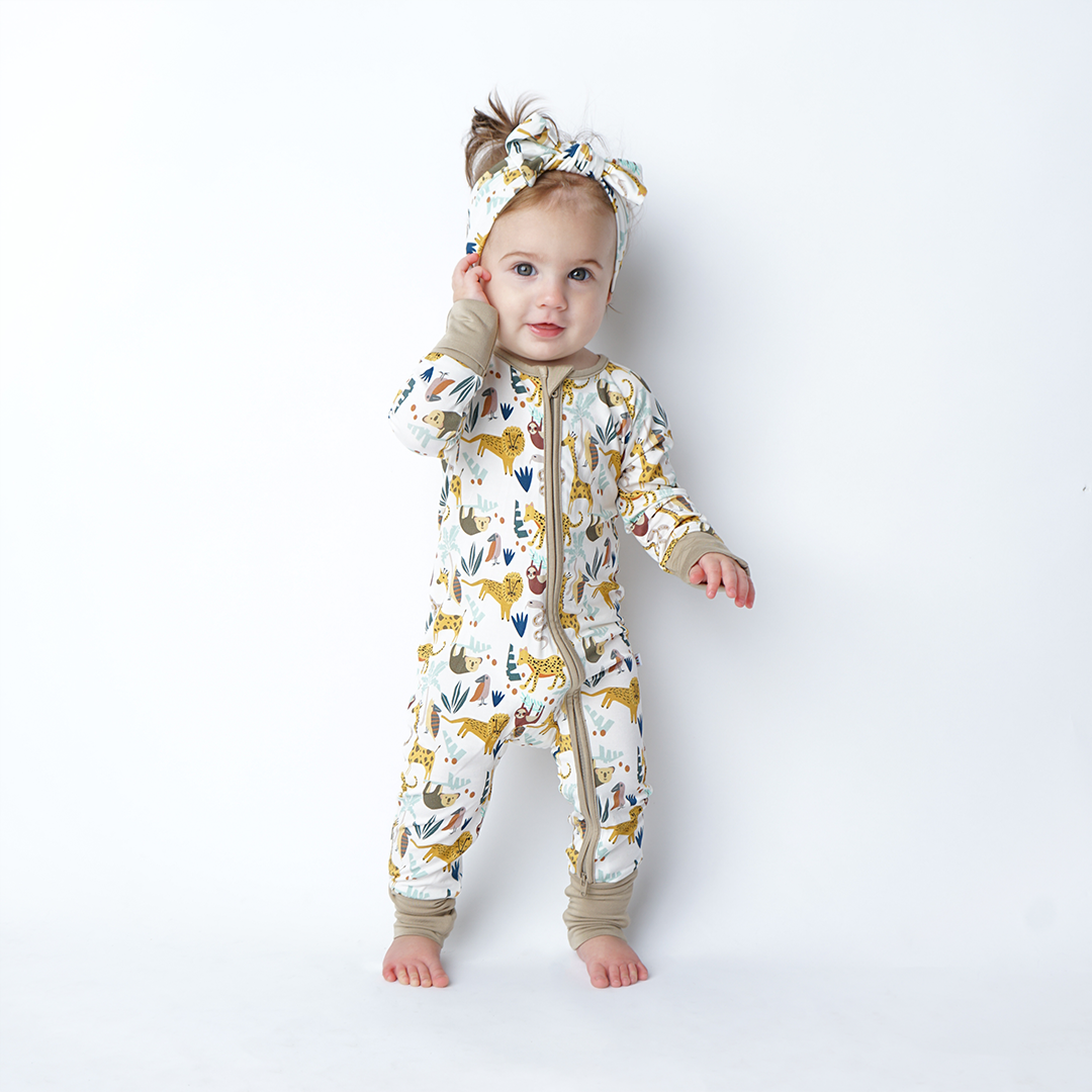 a baby stands in her "jungle friends" convertible. she is also wearing the headband. the "jungle friends" print is a mix of jungle animals that include giraffes, lions, cheetahs, sloths, snakes, and forest plants. 