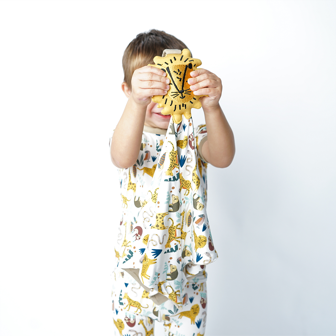 a young boy guards his face with the "jungle friends" lion lovey. the boy wears the 2-piece matching pajama set. the "jungle friends" print is a mix of jungle animals that include giraffes, lions, cheetahs, sloths, snakes, and forest plants. 