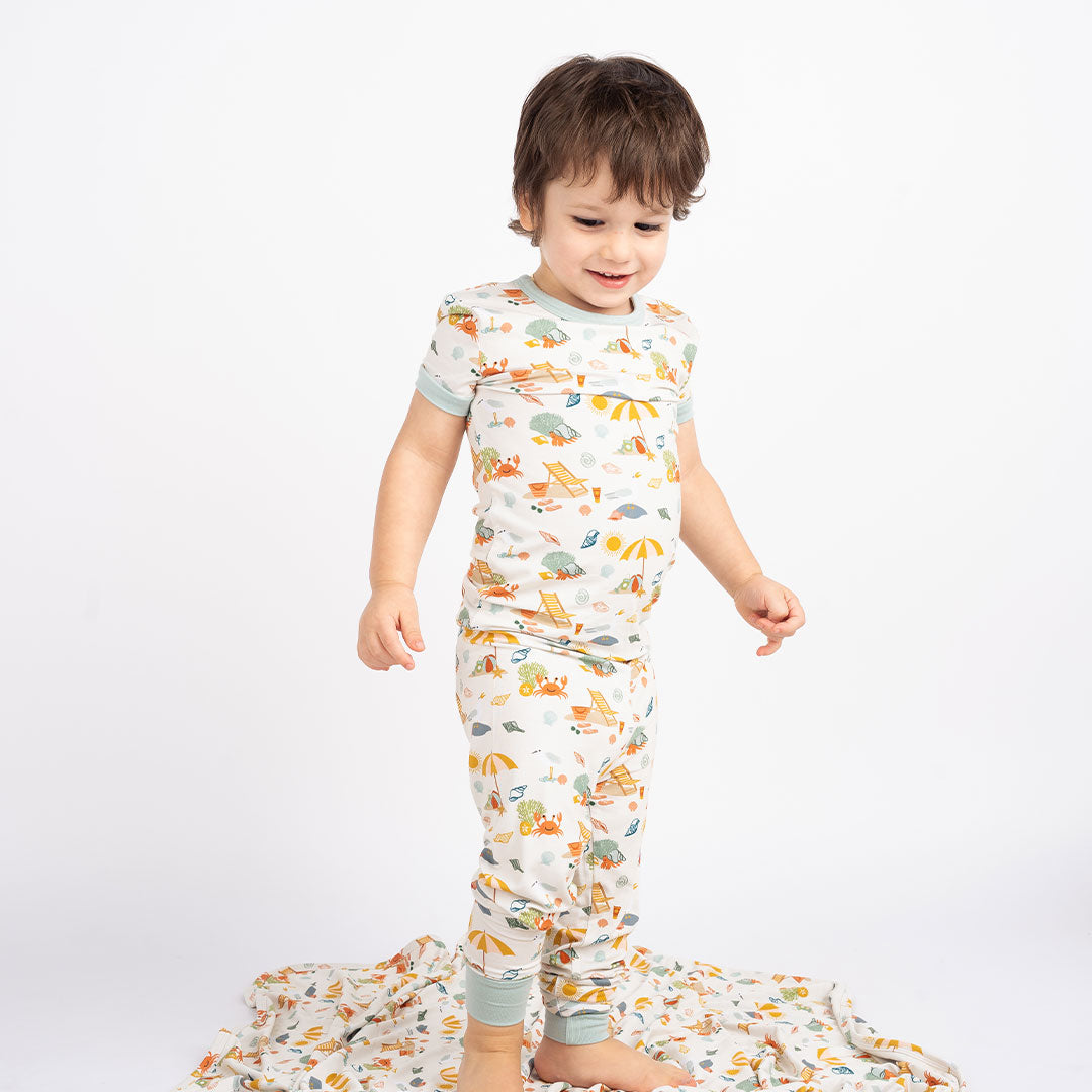 a young boy stands on the "beach days" bamboo blanket. he is wearing the "beach days" 2-piece matching pajama set. the "beach days" print is a scattered print of beach umbrellas, beach balls, sand buckets and shovels, coral, shells, beach chairs, seagulls, flipflops, and sunglass mixed in a orderly and creative way. 