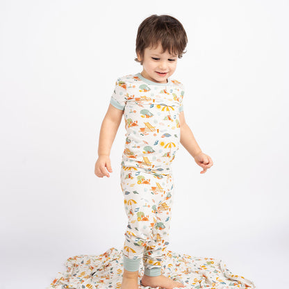 a young boy stands on the "beach days" bamboo blanket. he is wearing the "beach days" 2-piece matching pajama set. the "beach days" print is a scattered print of beach umbrellas, beach balls, sand buckets and shovels, coral, shells, beach chairs, seagulls, flipflops, and sunglass mixed in a orderly and creative way. 