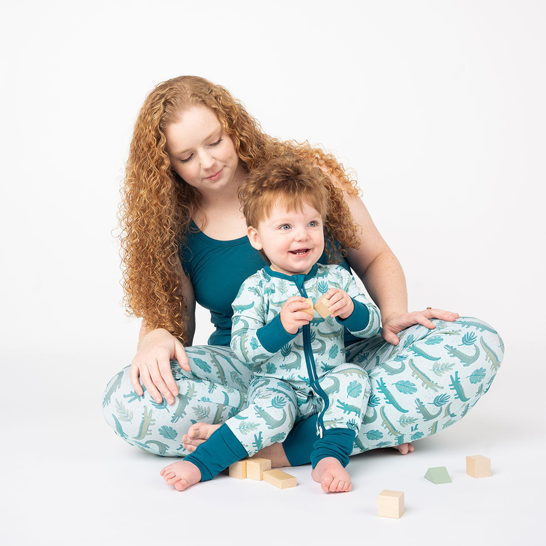 mother with her son in her lap. they are playing with wooden blocked, both in their "later gator" apparel. the "later gator" print has a mix of light and dark green alligators, leaves, and white dots scattered on a teal colored background. 