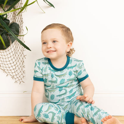 little boy sitting on the floor wearing his "later gator" matching pajama 2-piece set. the "later gator" print has a mix of light and dark green alligators, leaves, and white dots scattered on a teal colored background. 