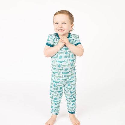boy smiling in his "later gator" 2-piece matching pajama set. the "later gator" print has a mix of light and dark green alligators, leaves, and white dots scattered on a teal colored background. 