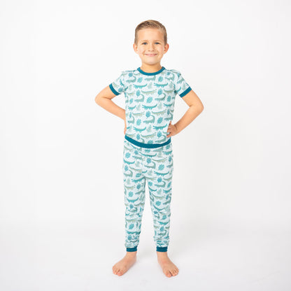 young boy posing in his "later gator" matching 2-piece pajama set. the "later gator" print has a mix of light and dark green alligators, leaves, and white dots scattered on a teal colored background. 