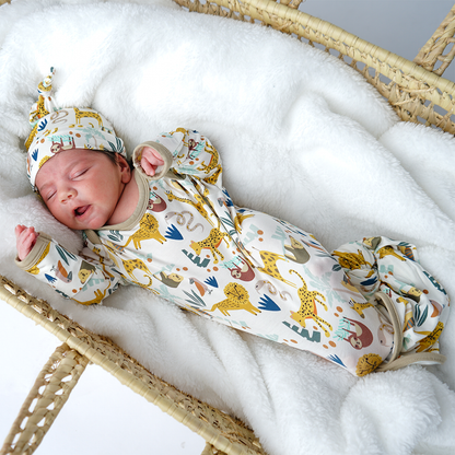 a baby lays in a basket cradle, on a white blanket. the baby wears the "jungle friends" matching gown and hat set. the "jungle friends" print is a mix of jungle animals that include giraffes, lions, cheetahs, sloths, snakes, and forest plants. 