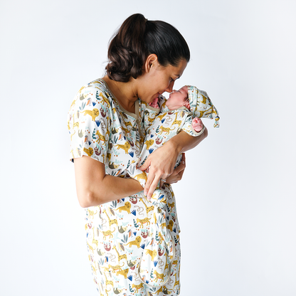 a mom touches noses with her newborn baby. they are wearing matching "jungle friends" apparel. the baby in the gown and hat set and the mom in the women's top and women's jogger pants. the "jungle friends" print is a mix of jungle animals that include giraffes, lions, cheetahs, sloths, snakes, and forest plants. 