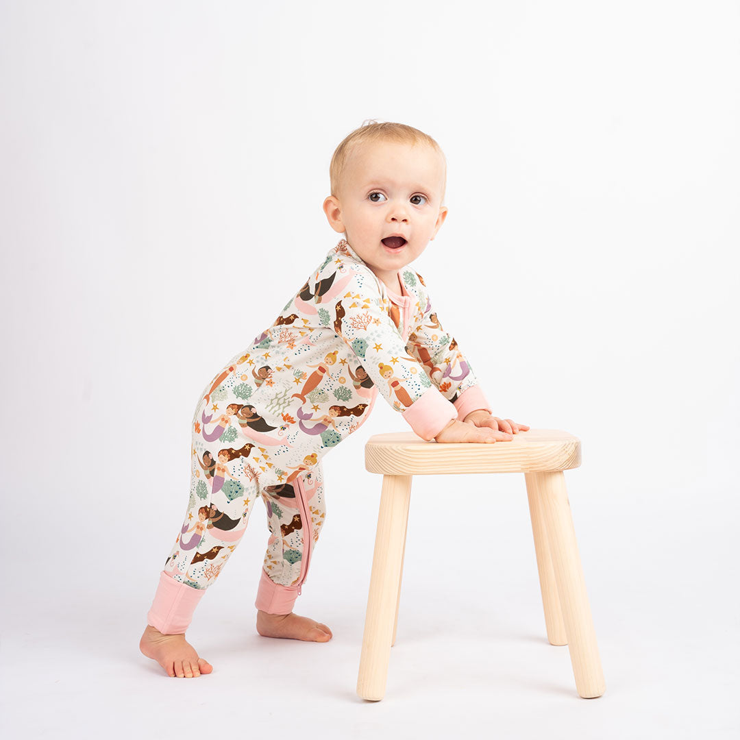 baby stands on their feet, using a wooden stool for balance. the baby is wearing the "making waves" convertible. the "making waves" print has a diverse spread of mermaids, sea coral, starfish, fish, and bubbles all spread out in different colors. this is all put on a beige background.