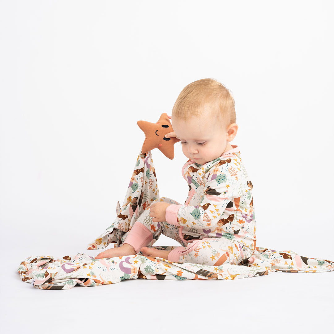 baby sits on the "making waves" bamboo blanket. the baby is holding up the "making waves" lovey and wears the "making waves" convertible. the "making waves" print has a diverse spread of mermaids, sea coral, starfish, fish, and bubbles all spread out in different colors. this is all put on a beige background.