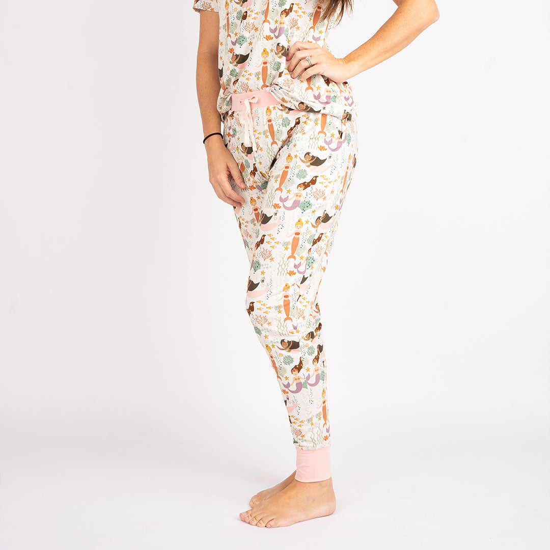 woman poses in the "making waves" women's top and the joggers. the "making waves" print has a diverse spread of mermaids, sea coral, starfish, fish, and bubbles all spread out in different colors. this is all put on a beige background.