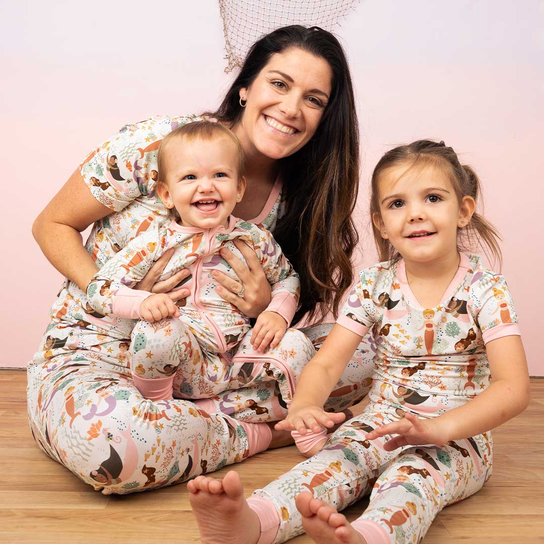 mom sits on the floor with her 2 kids. the baby is in the "making waves convertible. the older daughter is in the "making waves" 2-piece matching pajama set. the mom sits with the "making waves" women's top and the women's joggers. the "making waves" print has a diverse spread of mermaids, sea coral, starfish, fish, and bubbles all spread out in different colors. this is all put on a beige background.