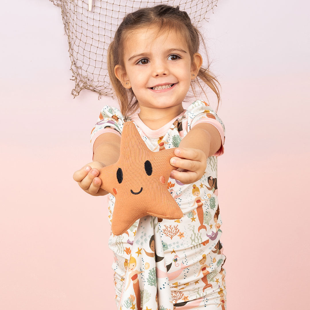 a girl smiles holding the "making waves" starfish lovey out for the camera. she wears the "making waves" 2-piece matching pajama set. she is standing in front of a pink ombre background that has a fish net on it. the "making waves" print has a diverse spread of mermaids, sea coral, starfish, fish, and bubbles all spread out in different colors. this is all put on a beige background.