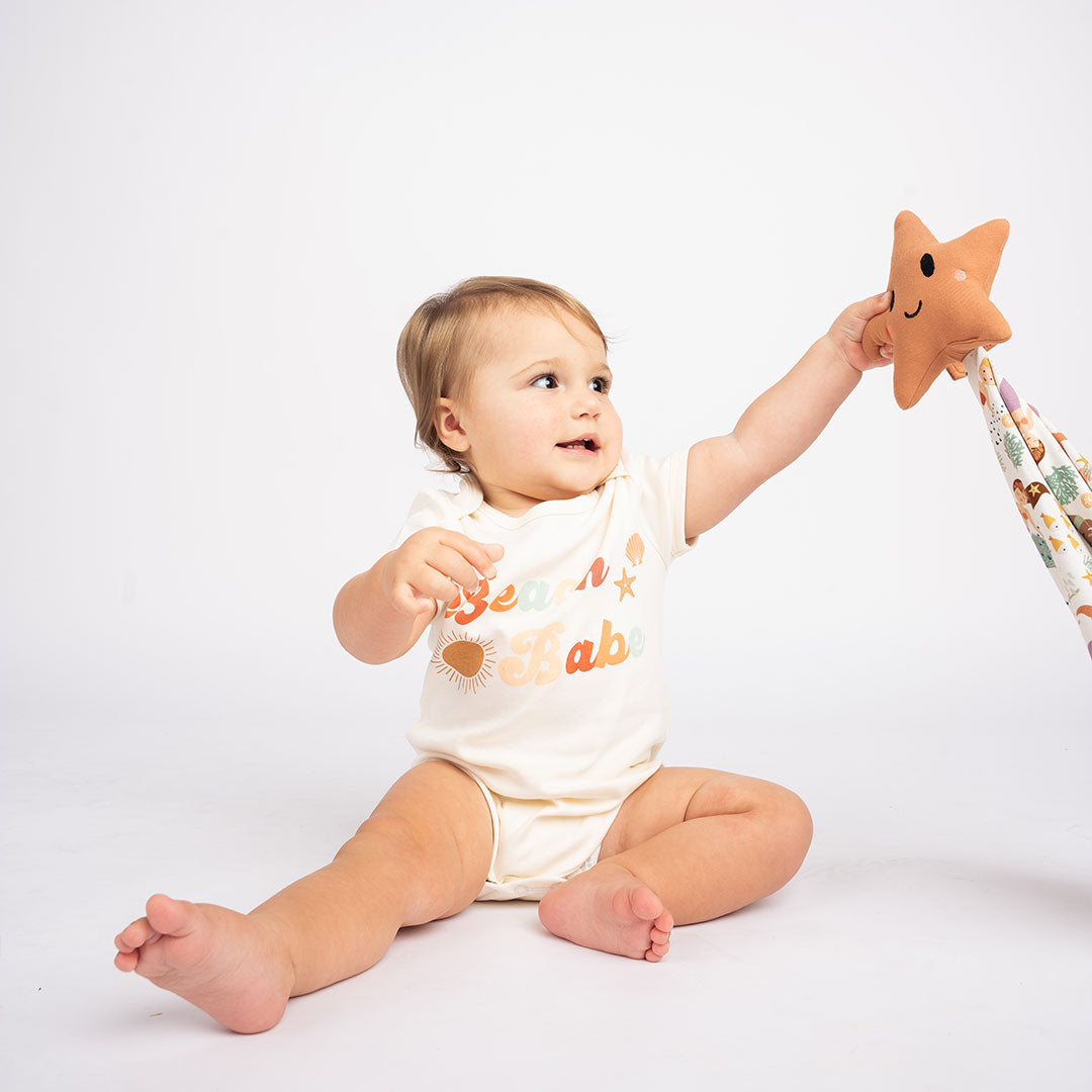 a baby sits on the floor in her "beach babe" onesie. she is holding the "making waves" starfish lovey. the "making waves" print has a diverse spread of mermaids, sea coral, starfish, fish, and bubbles all spread out in different colors. this is all put on a beige background.