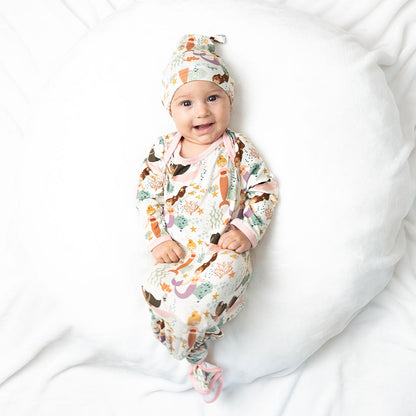 a smiling and happy baby laying on a soft white blanket. the baby is wearing the "making waves" gown and hat. the gown is tied in a knot at the bottom. the "making waves" print has a diverse spread of mermaids, sea coral, starfish, fish, and bubbles all spread out in different colors. this is all put on a beige background.