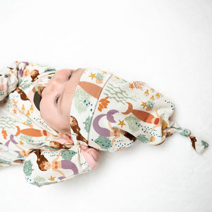 a picture of a baby laying on their back in the "making waves" gown and hat set. the "making waves" print has a diverse spread of mermaids, sea coral, starfish, fish, and bubbles all spread out in different colors. this is all put on a beige background.