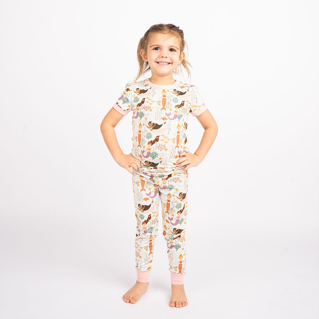 young girl posing with her hands on her hips. she's wearing the"making waves" short sleeved 2-piece pajama set. the "making waves" print has a diverse spread of mermaids, sea coral, starfish, fish, and bubbles all spread out in different colors. this is all put on a beige background.