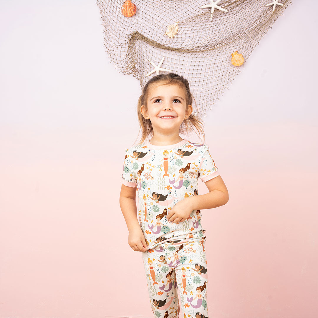 young girl poses, smiling in front of a pink ombre background. there's a fishnet with starfish and shells hanging behind her. she is wearing the "making waves" 2-piece matching pajama set. the "making waves" print has a diverse spread of mermaids, sea coral, starfish, fish, and bubbles all spread out in different colors. this is all put on a beige background.