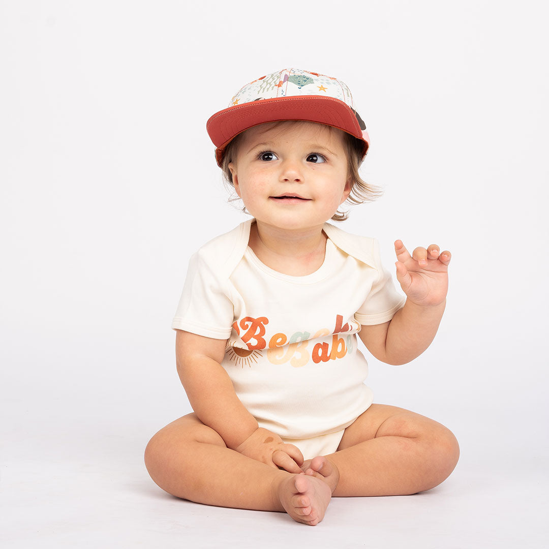 a baby sits on the floor with his hand in the air. he is wearing the "making waves" snapback hat and the "beach babe" onesie. the "making waves" print has a diverse spread of mermaids, sea coral, starfish, fish, and bubbles all spread out in different colors. this is all put on a beige background.